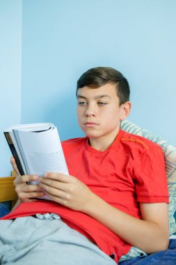 Beyaz genç çocuk yatakta oturarak kitap okuma