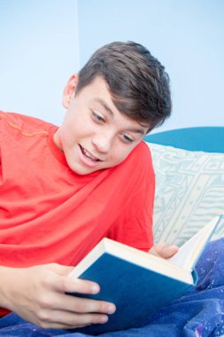 Beyaz genç çocuk bir yatak gülüyor döşeme bir kitap okuma