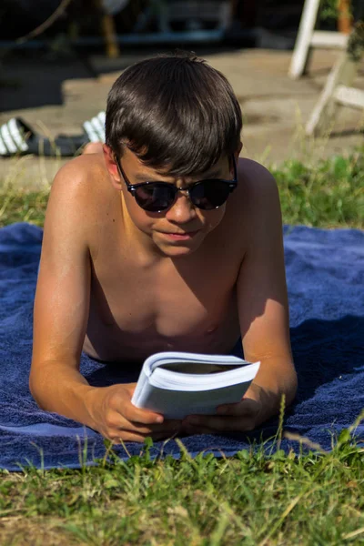 十几岁的男孩在花园里晒太阳时看书 戴着墨镜 — 图库照片
