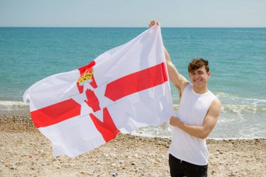 Caucasian male on a beach holding an Ulster flag clipart