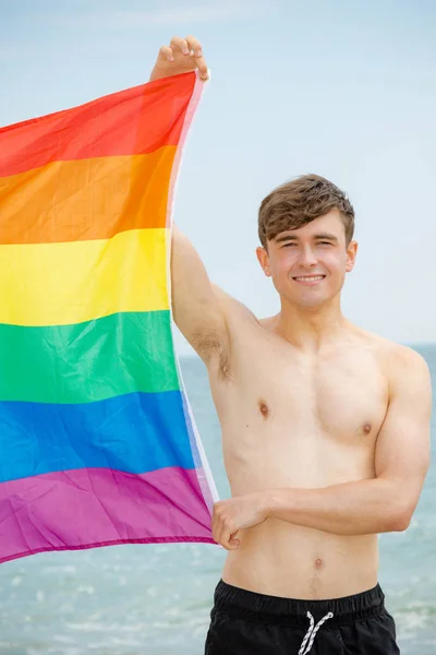 Caucasian male on a beach holding a Pride flag