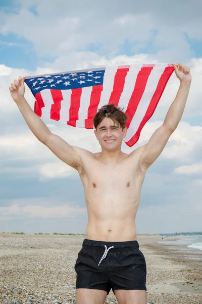 Homem caucasiano em uma praia segurando uma bandeira americana — Fotografia de Stock