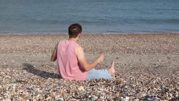 彼のシャツを脱ぐビーチに座っている若いコーカサスの成人男性 — ストック動画