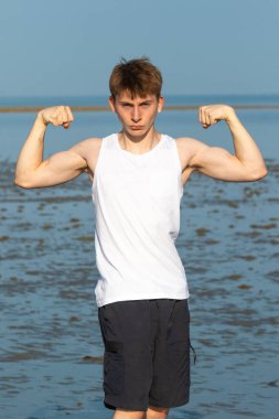 Genç çocuk kol kaslarını esnetiyor kolsuz bir bluz ve kumsalda şort giyiyor.