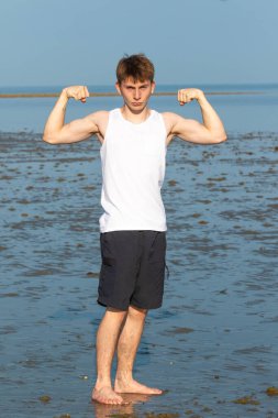 Genç çocuk kol kaslarını esnetiyor kolsuz bir bluz ve kumsalda şort giyiyor.