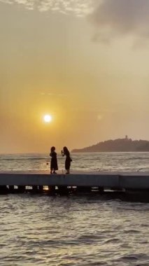 Kadın fotoğrafçı, Audin, Kusadasi 'de yaz akşamı gökyüzünde dalgalanan deniz ve gün batımına karşı iskelede siyahlı kadının fotoğraflarını çekiyor. Yüksek kalite 4k görüntü