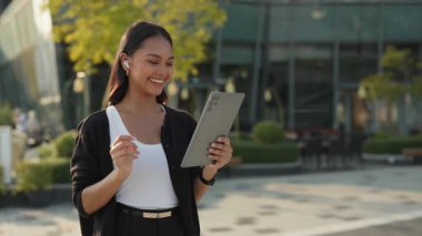Asyalı mutlu iş kadınının portresi şehirdeki dijital tabletle arkadaşlarıyla görüntülü görüşme yapıyor. Kadın, şehir merkezinde mutluluk ve profesyonelliği ifade eden çevrimiçi toplantıya katıldı