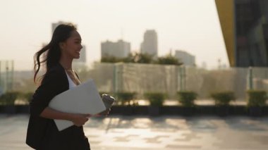 Kahveli mutlu bir Asyalı kadının portresi ve günbatımı caddesinde dizüstü bilgisayarla yürüyor. Pozitif tavır sergileyen kadın girişimci, şehirde bilgisayar ve kupa taşıyor.