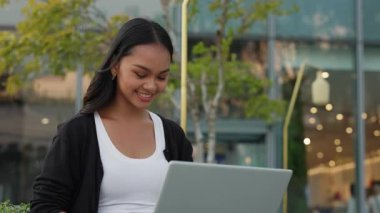 Gülümseyen Asyalı kadın yöneticinin portresi dizüstü bilgisayar kullanıyor ve şirket binasının yanında oturuyor. Şirin bayan girişimci açık havada bilgisayar aracılığıyla müşterilerle çevrimiçi iletişim kuruyor.