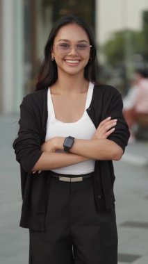 Gözlüklü pozitif Asyalı kadın portresi şehir merkezinde kollarını kavuşturuyor. Genç esmer kadın ayakta duruyor ve şehirde dikey çekimde profesyonelliğini gösteriyor. Şirket çalışanı