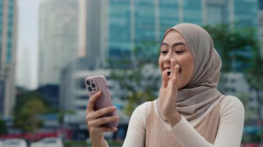 Mutlu Müslüman kadın portresi gökdelenlere karşı akıllı telefon standlarında görüntülü sohbet kullanıyor. Genç Malezyalı kadın, şehirdeki video dalgalarına cevap veriyor. Çevrimiçi iletişim