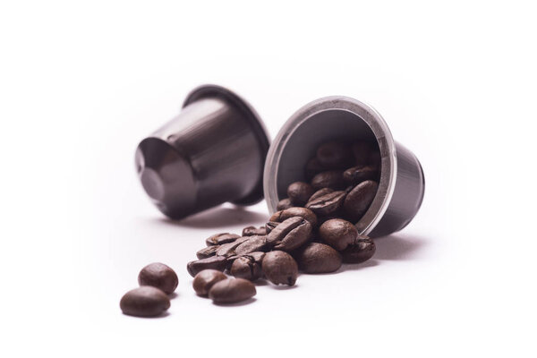 Toasted coffee beans spill out from a capsule on white background
