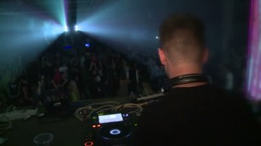 Tekno parti. İnsanlar dans eder, DJ çalar. Her yerde renkli ışıklar