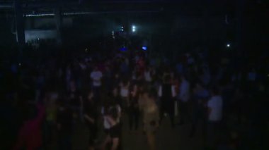 Tekno parti. İnsanlar dans eder, DJ çalar. Her yerde renkli ışıklar