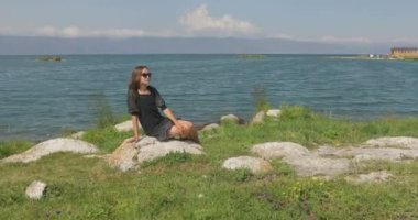 Kız rıhtımda takılıyor, sahilde yürüyor. Kameranın önünde poz vermek, gülümsemek, kumda yalın ayak yürümek, rüzgarın şalla oynaması.
