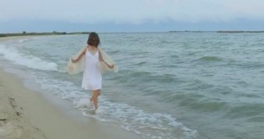 Kumsalda yürüyen bir kız. Kameranın önünde poz vermek. Gülümsüyor, kumda yalın ayak yürüyor, rüzgar şalla çalınıyor.