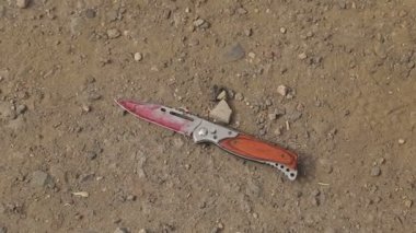 Bir adam yerden bir bıçak alıyor, kırmızı bir mendille bıçağın üzerindeki kan lekelerini çıkarıyor, açık havada, bıçaktaki kan izlerini siliyor ve arabada sarı bir bez ile temizliyor.