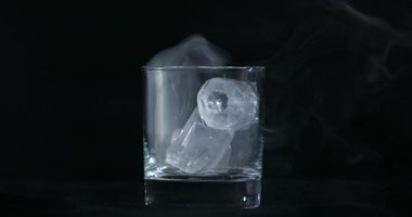 Buz küpleri alkolde eriyor, viski bardağı ve duman siyah arka planda yüzüyor.