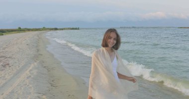 Kumsalda yürüyen bir kız. Kameranın önünde poz vermek. Gülümsüyor, kumda yalın ayak yürüyor, rüzgar şalla çalınıyor.