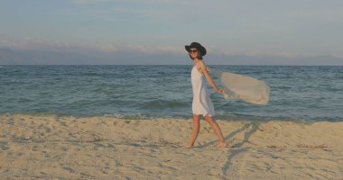 Kumsalda yürüyen bir kız. Kameranın önünde poz vermek. Gülümsüyor, kumda yalın ayak yürüyor, rüzgar şalla çalınıyor.