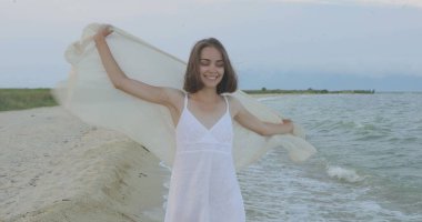 Kumsalda yürüyen bir kız. Kameranın önünde poz vermek. Gülümsüyor, kumda yalın ayak yürüyor, rüzgar şalla çalınıyor.