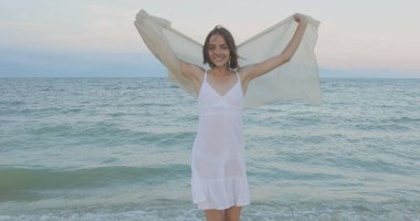 Kumsalda yürüyen bir kız. Kameranın önünde poz vermek. Gülümsüyor, kumda yalın ayak yürüyor, rüzgar şalla çalınıyor.