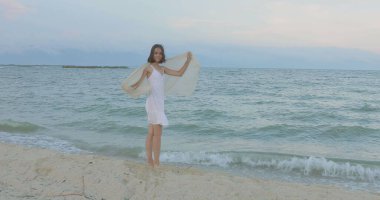 Kumsalda yürüyen bir kız. Kameranın önünde poz vermek. Gülümsüyor, kumda yalın ayak yürüyor, rüzgar şalla çalınıyor.