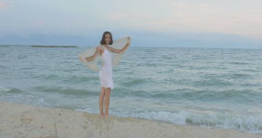 Kumsalda yürüyen bir kız. Kameranın önünde poz vermek. Gülümsüyor, kumda yalın ayak yürüyor, rüzgar şalla çalınıyor.