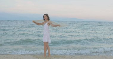 Kumsalda yürüyen bir kız. Kameranın önünde poz vermek. Gülümsüyor, kumda yalın ayak yürüyor, rüzgar şalla çalınıyor.