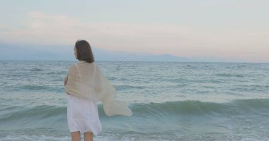 Kumsalda yürüyen bir kız. Kameranın önünde poz vermek. Gülümsüyor, kumda yalın ayak yürüyor, rüzgar şalla çalınıyor.