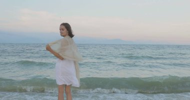 Kumsalda yürüyen bir kız. Kameranın önünde poz vermek. Gülümsüyor, kumda yalın ayak yürüyor, rüzgar şalla çalınıyor.