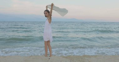 Kumsalda yürüyen bir kız. Kameranın önünde poz vermek. Gülümsüyor, kumda yalın ayak yürüyor, rüzgar şalla çalınıyor.