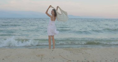 Kumsalda yürüyen bir kız. Kameranın önünde poz vermek. Gülümsüyor, kumda yalın ayak yürüyor, rüzgar şalla çalınıyor.