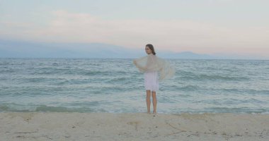 Kumsalda yürüyen bir kız. Kameranın önünde poz vermek. Gülümsüyor, kumda yalın ayak yürüyor, rüzgar şalla çalınıyor.