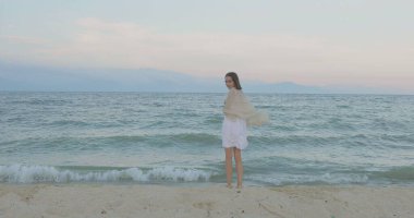 Kumsalda yürüyen bir kız. Kameranın önünde poz vermek. Gülümsüyor, kumda yalın ayak yürüyor, rüzgar şalla çalınıyor.