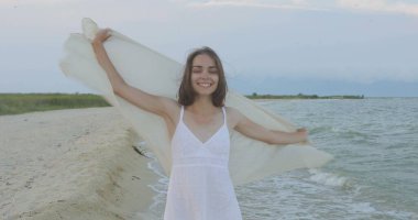 Kumsalda yürüyen bir kız. Kameranın önünde poz vermek. Gülümsüyor, kumda yalın ayak yürüyor, rüzgar şalla çalınıyor.