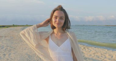 Kumsalda yürüyen bir kız. Kameranın önünde poz vermek. Gülümsüyor, kumda yalın ayak yürüyor, rüzgar şalla çalınıyor.