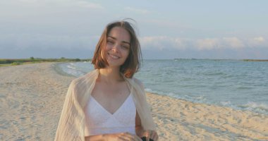 Kumsalda yürüyen bir kız. Kameranın önünde poz vermek. Gülümsüyor, kumda yalın ayak yürüyor, rüzgar şalla çalınıyor.