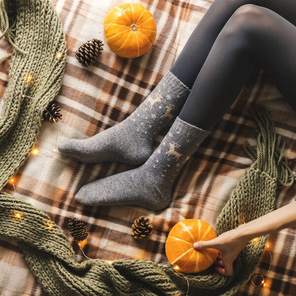 Womens hands and feet in sweater and woolen cozy gray socks sitting on plaid with pumpkin, pine cones, garland, knitted scarf and leaves. Concept winter comfort, morning drinking.