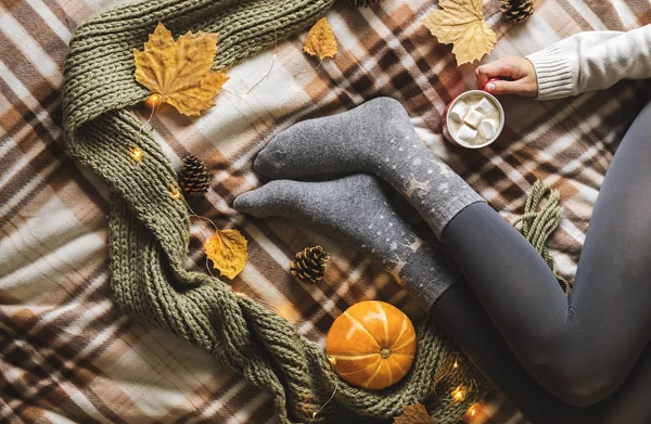 Womens hands and feet in sweater and woolen cozy gray socks holding cup of hot coffee with marshmallow, sitting on plaid with pumpkin, knitted scarf, leaves. Concept winter comfort, morning drinking. — Stock Photo, Image