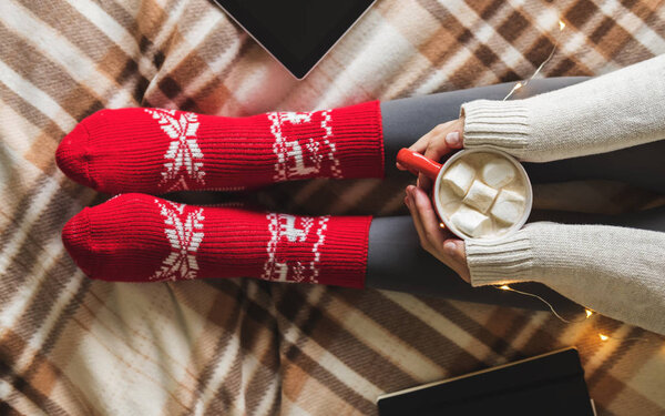 Womens hands and feet in sweater and woolen cozy red socks holding cup of hot coffee with marshmallow, sitting on plaid with garland, tablet and notebook. Concept winter comfort, morning drinking.