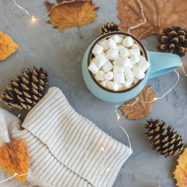 Tasse bleue avec café, chocolat chaud ou cacao avec guimauve sur fond de béton gris et feuilles sèches allongées, cônes, pull, guirlande. Concept d'automne ou d'hiver chaleureux et confortable . — Photo