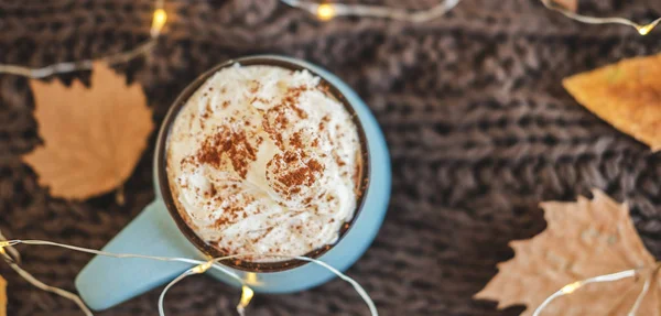 Mug of coffee, cocoa or hot chocolate with whipped cream and cinnamon on scarf with leaves, garland. Pumpkin latte - cozy drink for cold fall or winter. — Stock Photo, Image