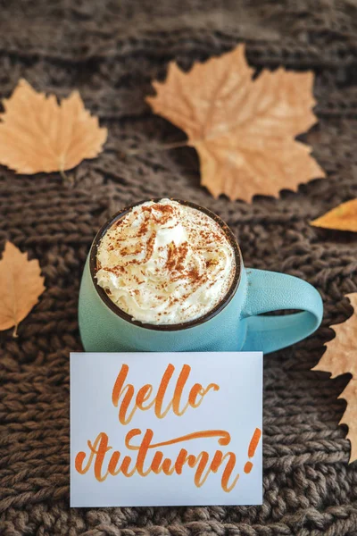 Mug of coffee, cocoa or hot chocolate with whipped cream and cinnamon on scarf with leaves, card Hello Autumn. Pumpkin latte - cozy drink for cold autumn or winter.