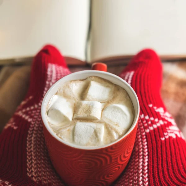 Женские ножки в красных вязаных носках держат чашку горячего кофе, шоколад, какао с зефиром на клетке. зимний комфорт, инвалидность, утреннее питье, тепло посреди холода . — стоковое фото
