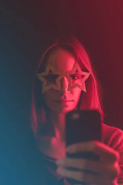 Girl neon light, dressed in glasses for party in form of stars, takes selfie on the phone.