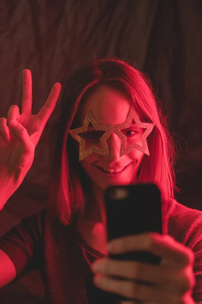 Girl neon light, dressed in glasses for party in form of stars, takes selfie on the phone.
