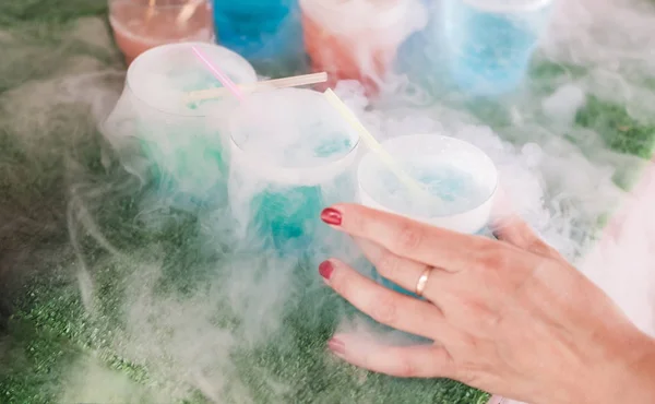 Steaming multi-colored cocktails with dry ice standing on green background. Female hand takes one cocktail. — Stock Photo, Image