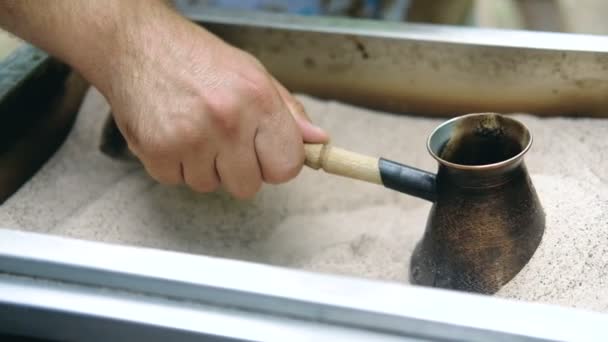 Mans hand håller Turk och förbereder turkiskt kaffe i sanden i den. Slow motion. — Stockvideo