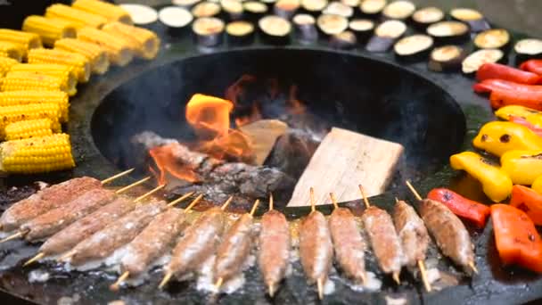 Barbecue rond avec feu ouvert à l'intérieur. Des repas pour le pique-nique d'été sont en préparation : maïs, aubergine, poivron, kebab. Les mains masculines en gants noirs retournent la nourriture avec des pinces pour barbecue . — Video